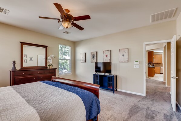 A bedroom with a bed, a ceiling fan, a dresser, a TV, and a door leading to a kitchen.
