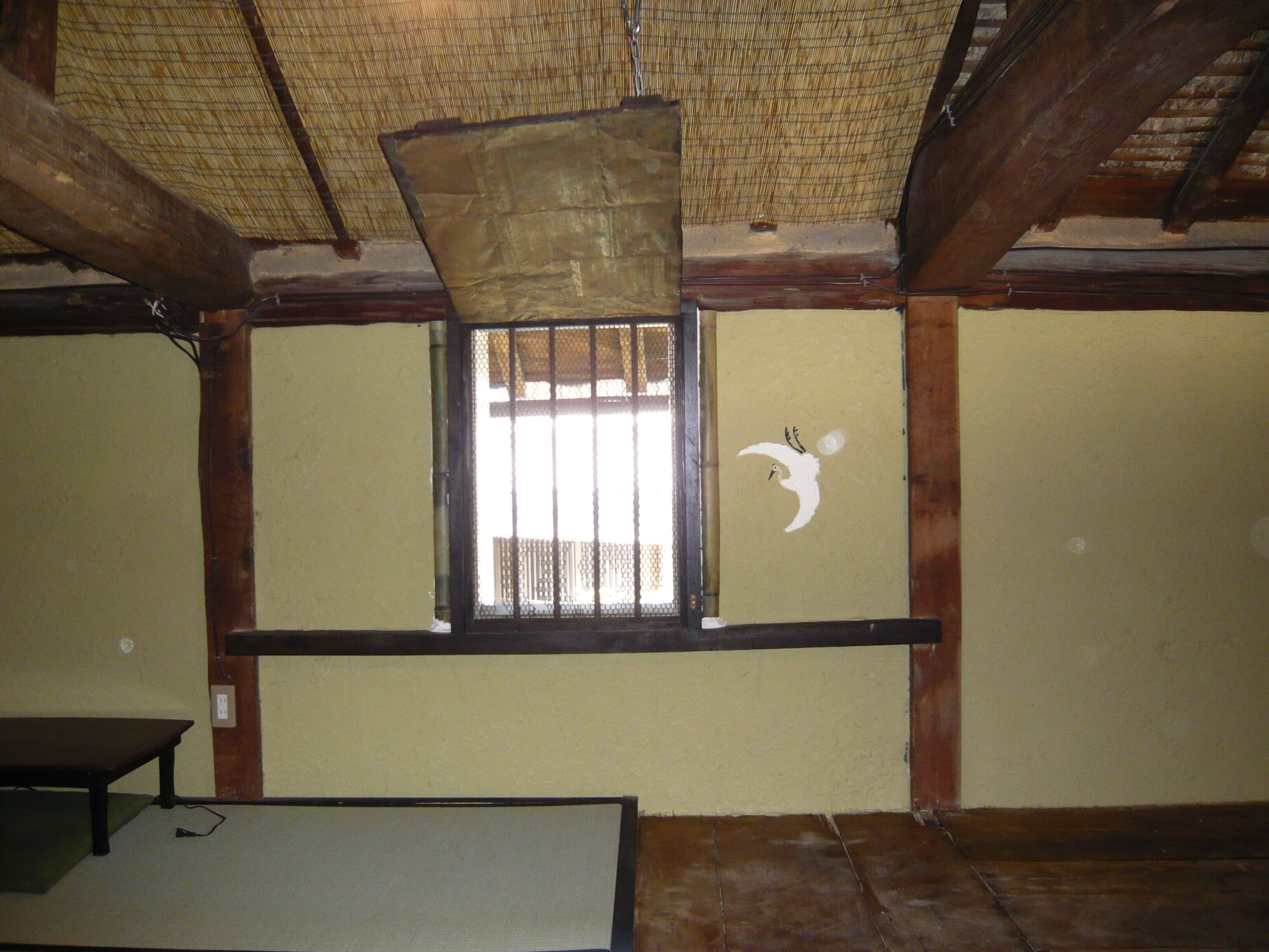 A room with a traditional wooden ceiling, a window with a bamboo blind, and a wall with a white bird mural.