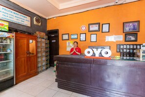 Ein Empfangsschalter mit einem Schild, auf dem 'OYO' steht, und eine Frau, die dahinter steht. An der Wand befinden sich gerahmte Zertifikate und Bilder, ein Kühlschrank mit Getränken und ein Holzkasten mit Kartons.
