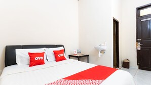 Une chambre d’hôtel avec un lit, deux oreillers rouges, une couverture blanche et rouge, une table de chevet avec une bouteille et des mouchoirs, un lavabo blanc et une porte.