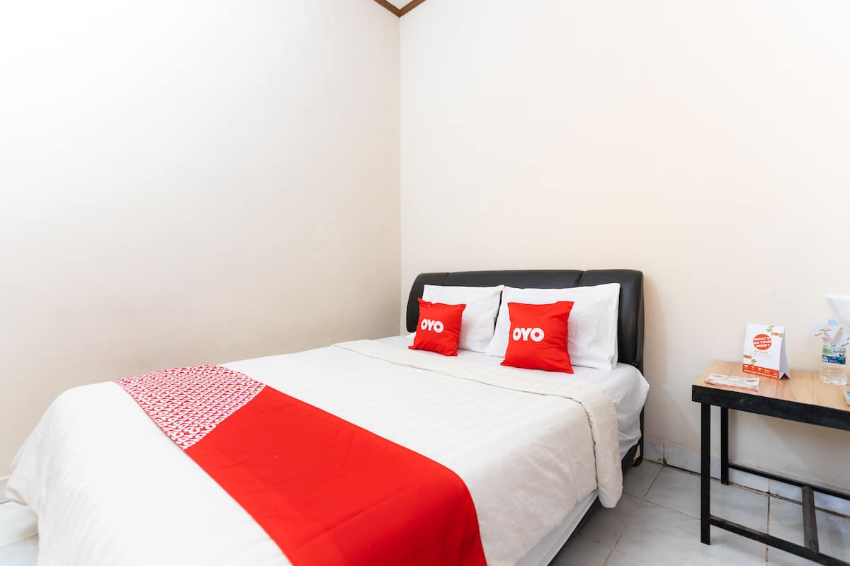 Habitación de hotel con cama, dos almohadas rojas, cubrecama blanco y una mesita con botella de agua y menú.