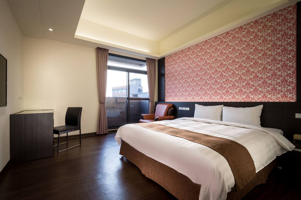 A hotel room with a large bed, a desk, a chair, and a window with curtains.