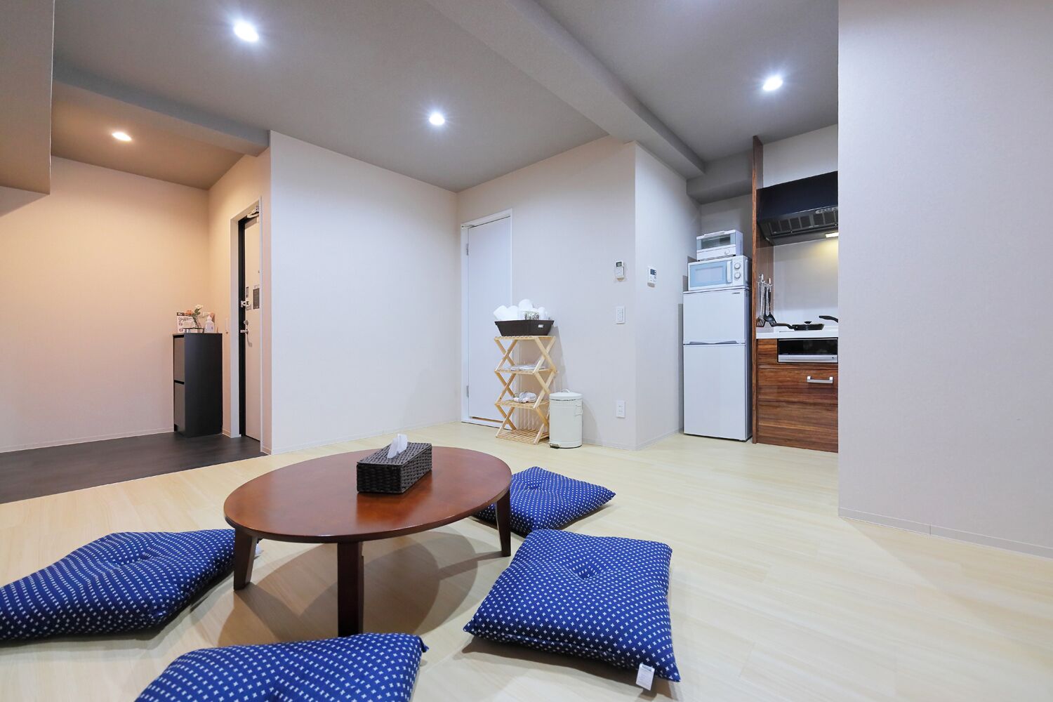 A compact living space with a round table and four blue pillows, a kitchenette, and a small trash bin.