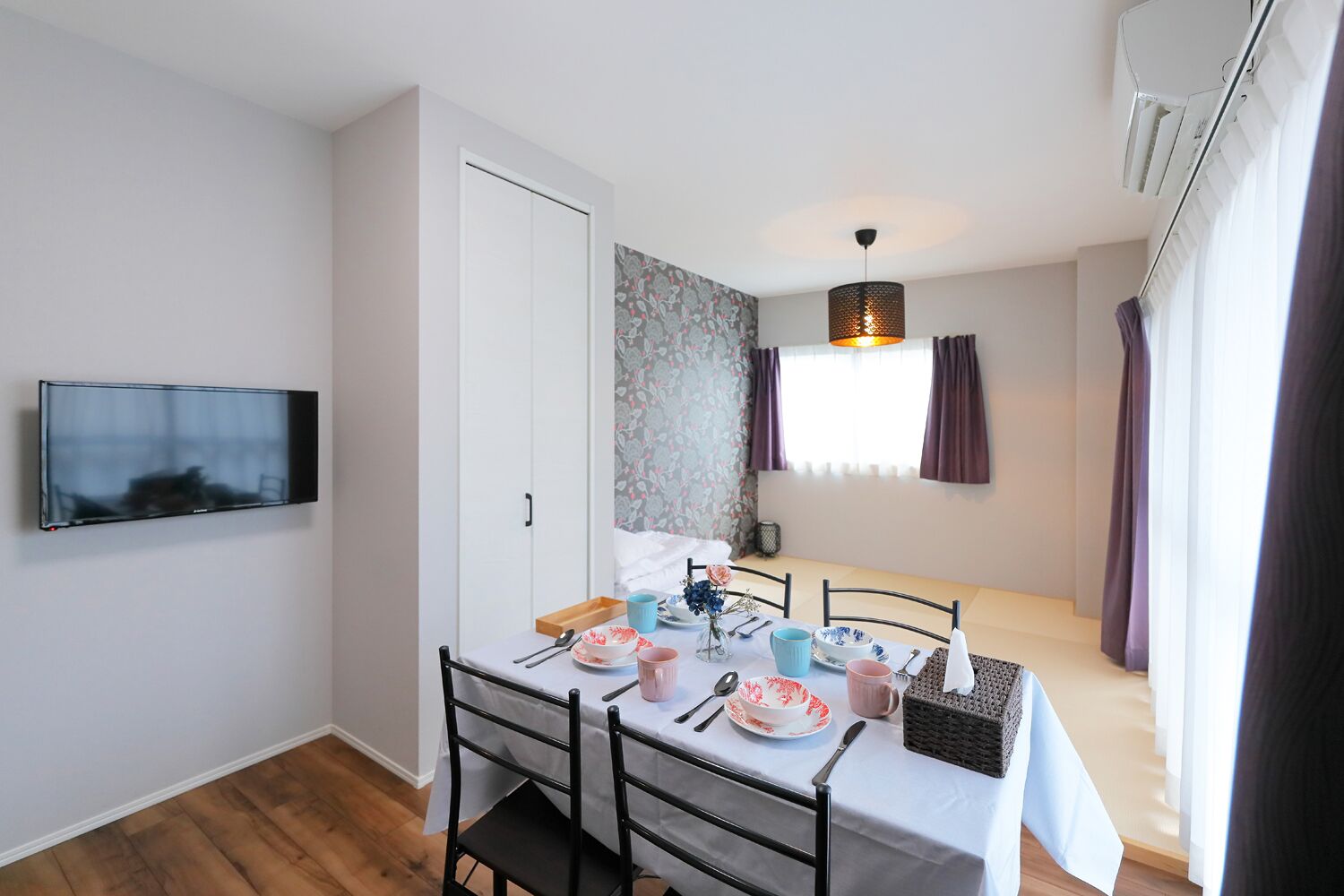 A dining area with a table set for four, a flat-screen TV mounted on the wall, and a window with curtains.