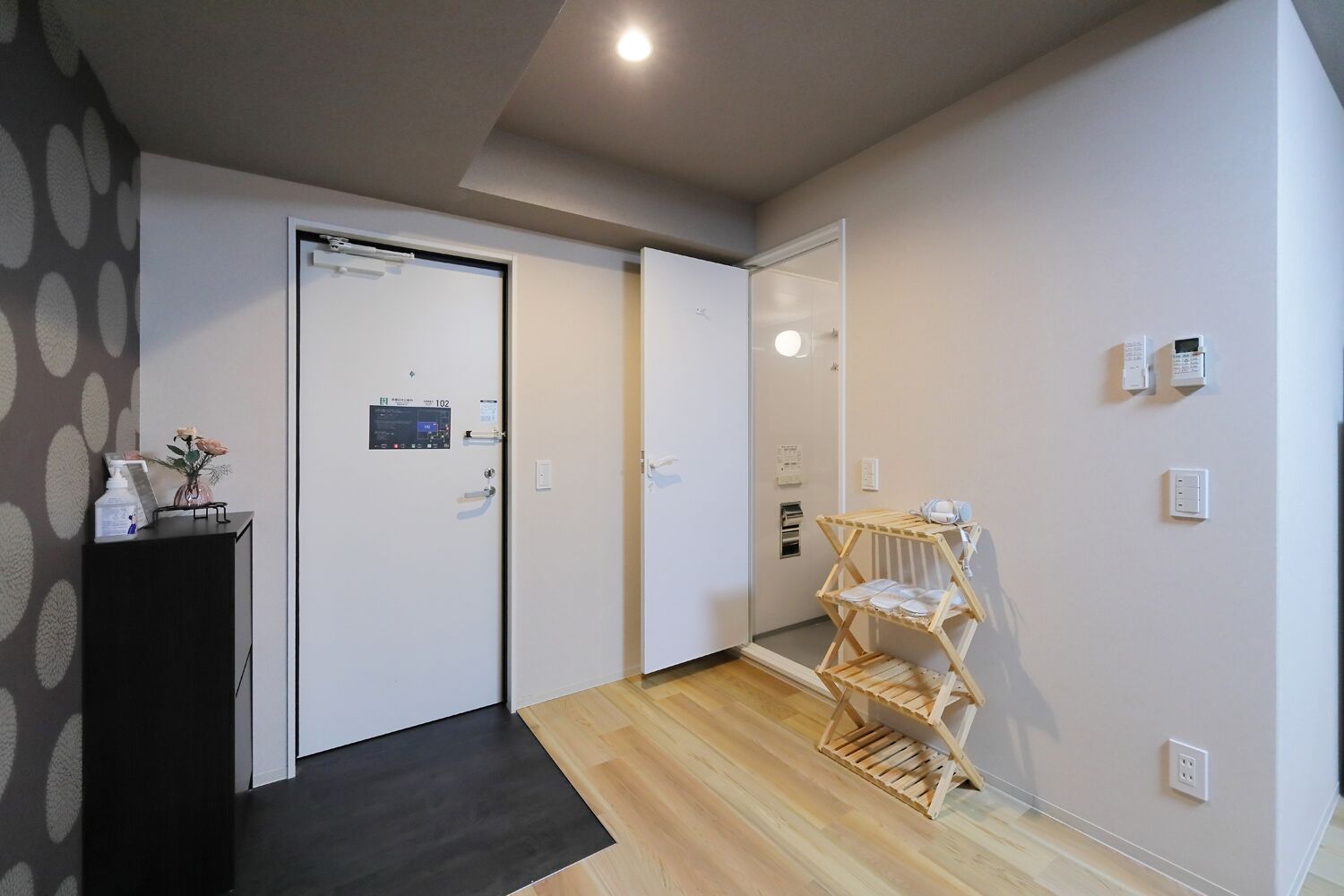A hallway with a wooden floor, a black cabinet with a vase of flowers, a door with a control panel, and a wooden rack with folded clothes.