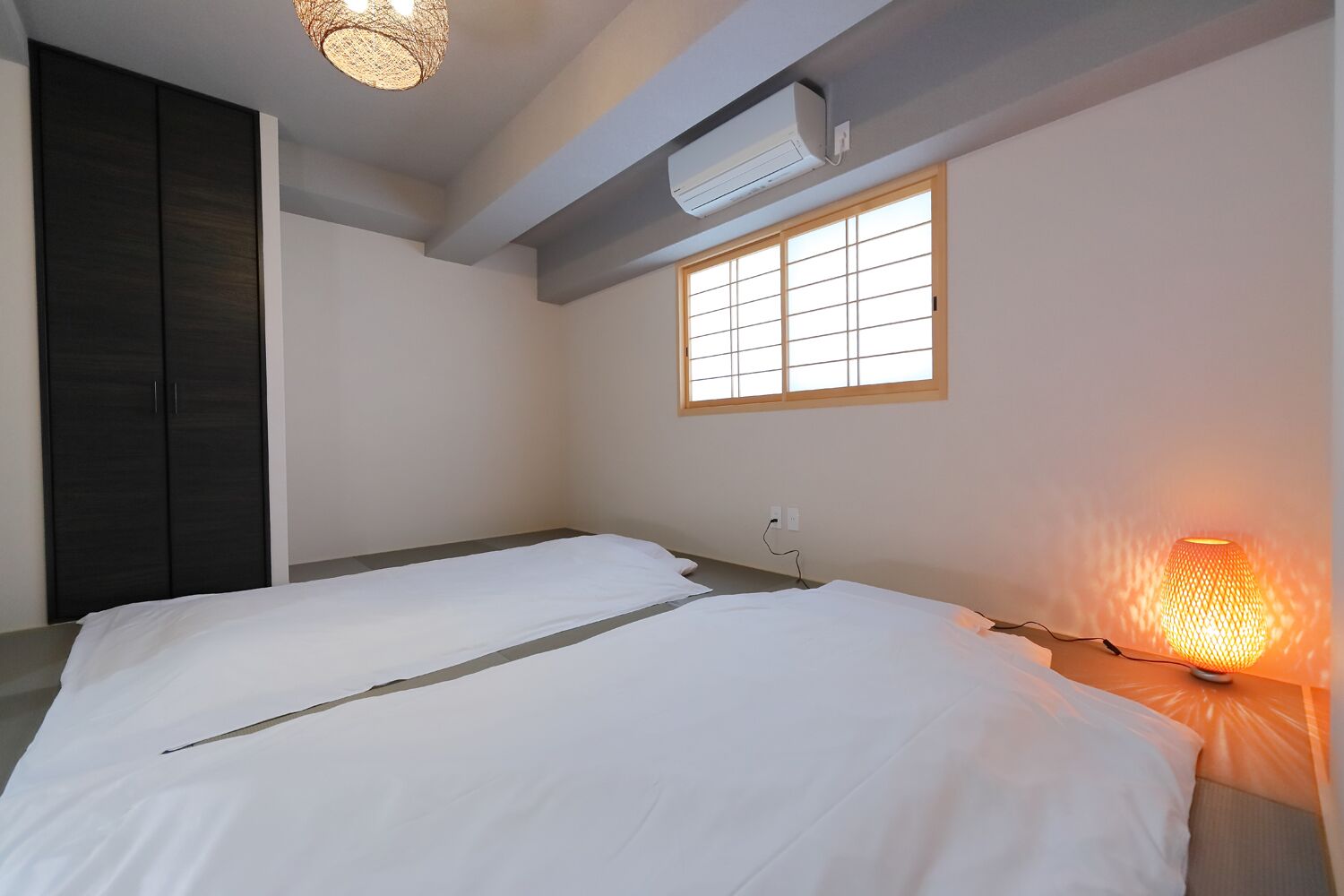 A minimalist room with two beds, a window with wooden frames, a wall-mounted air conditioner, and a hanging light fixture.
