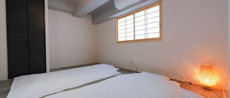 A minimalist room with two beds, a window with wooden frames, a wall-mounted air conditioner, and a hanging light fixture.