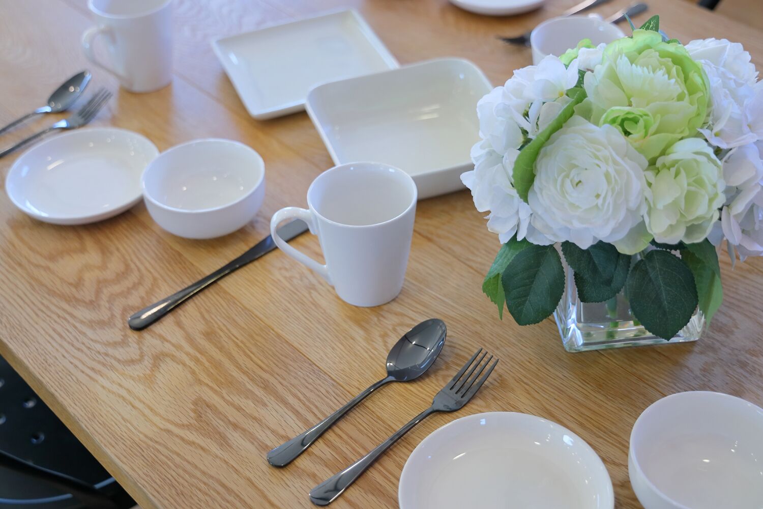 Una mesa de madera dispuesta con platos, fuentes, tazas y cubiertos, junto a un florero con flores blancas.