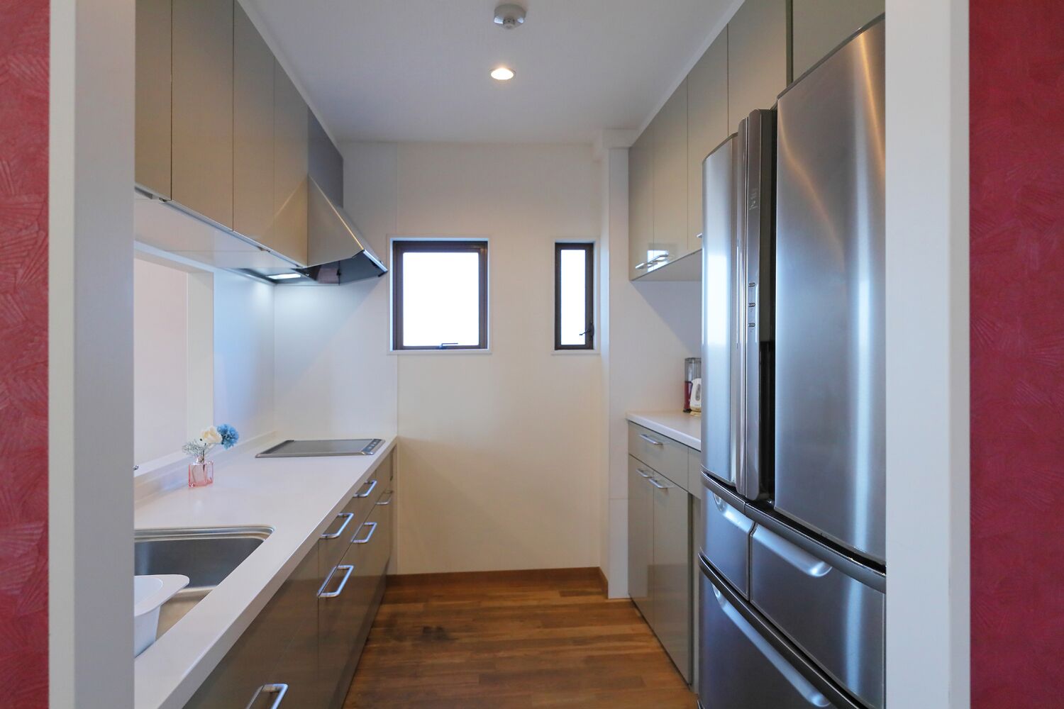 A narrow kitchen with a stainless steel refrigerator, a window, and a wooden floor.