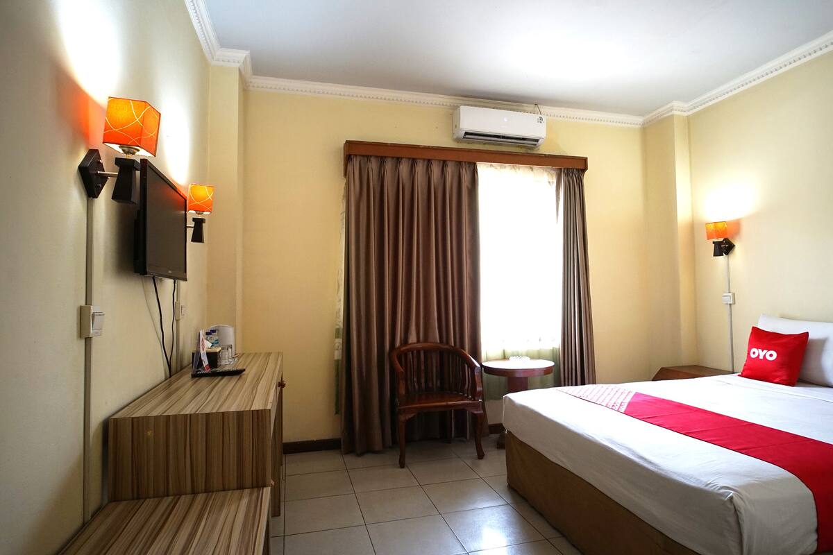 A hotel room with a bed, a desk, a chair, a small table, and an air conditioning unit.
