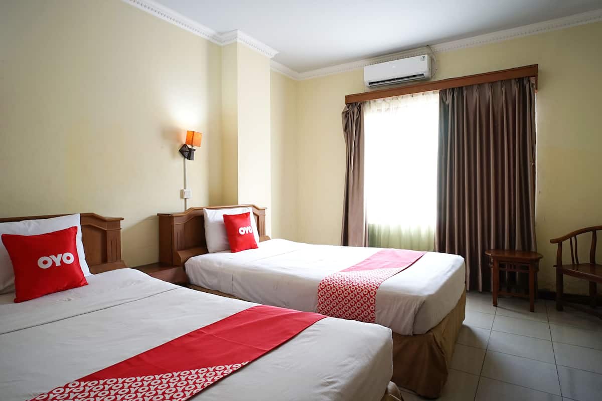 A hotel room with two beds, each with a red OYO pillow, a patterned red blanket, and a white bedspread. There is a wooden nightstand with a lamp, a small table, and a chair. An air conditioning unit is mounted on the wall above the window, which has brown curtains.