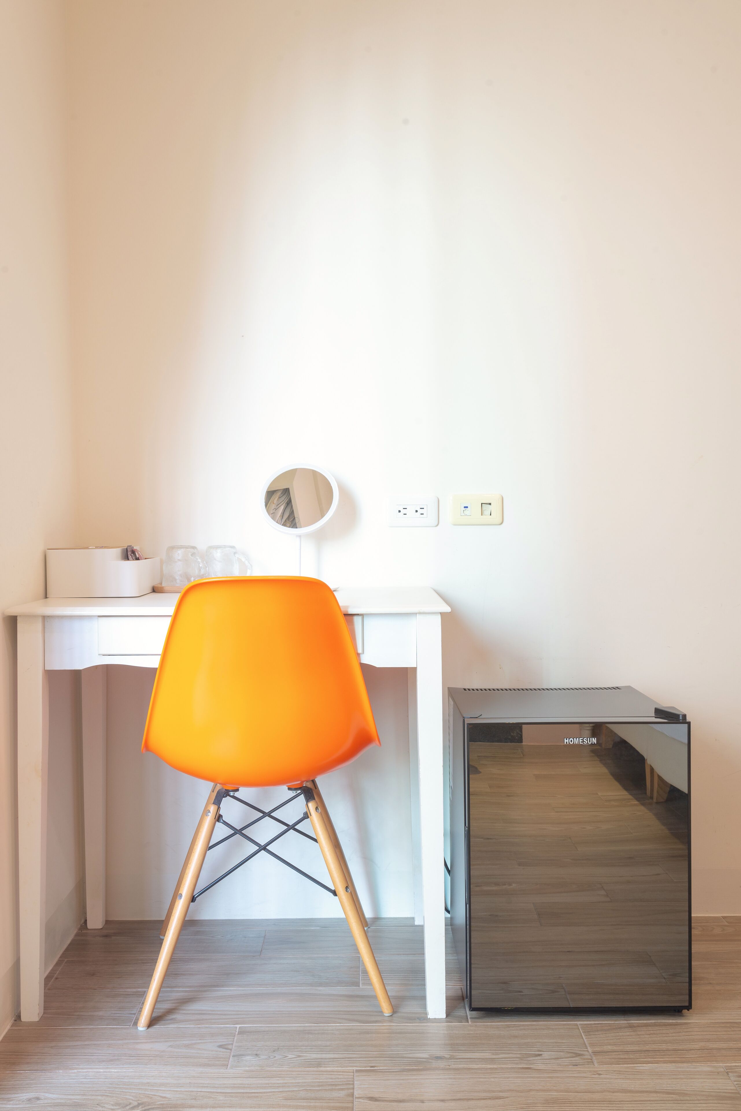 Une pièce de taille modeste, avec un bureau blanc, une chaise orange, un petit miroir et un meuble de rangement noir.