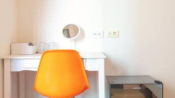 Une pièce de taille modeste, avec un bureau blanc, une chaise orange, un petit miroir et un meuble de rangement noir.
