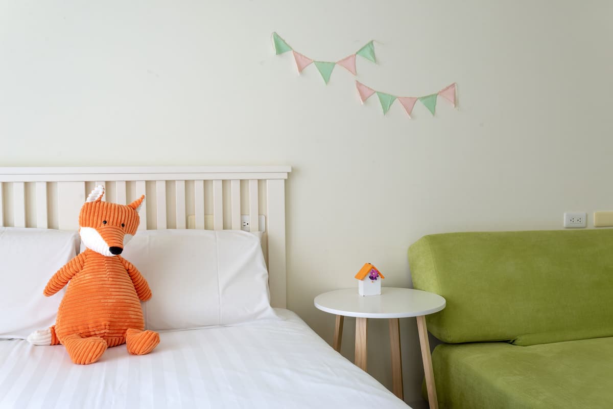 Una cama con un peluche, una mesita con un objeto con forma de casa y una silla verde.