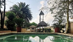 Una piscina con una estructura de techo de paja, rodeada de exuberante follaje y un pequeño edificio.
