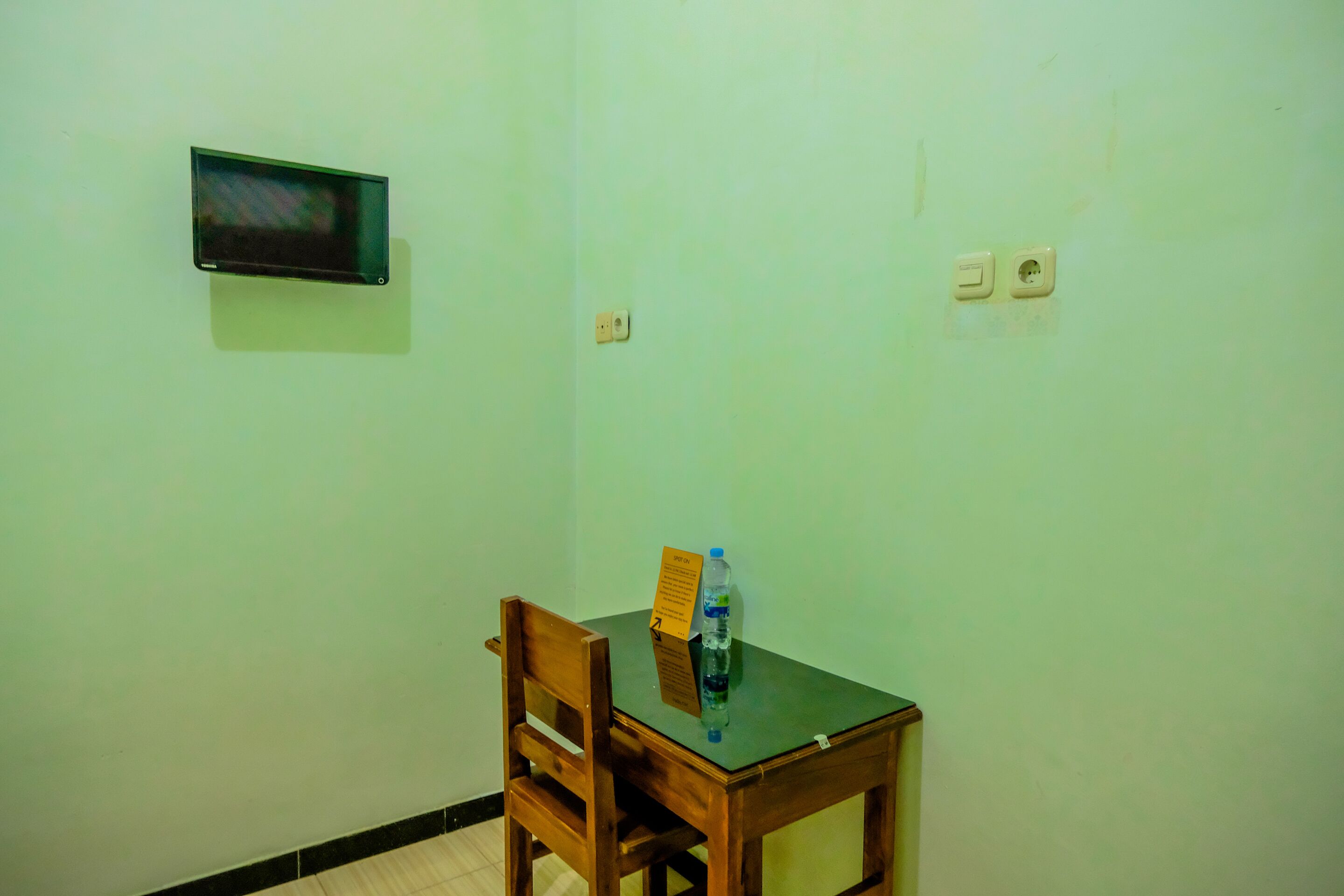 Une petite pièce avec une chaise en bois, une table, un téléviseur fixé au mur et une bouteille d’eau sur la table.
