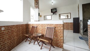 A room with a brick wall, a TV mounted on the wall, and a window. There are two wooden chairs and a small round table. - OYO 2021 Wisma Vandera Syariah (Garut)