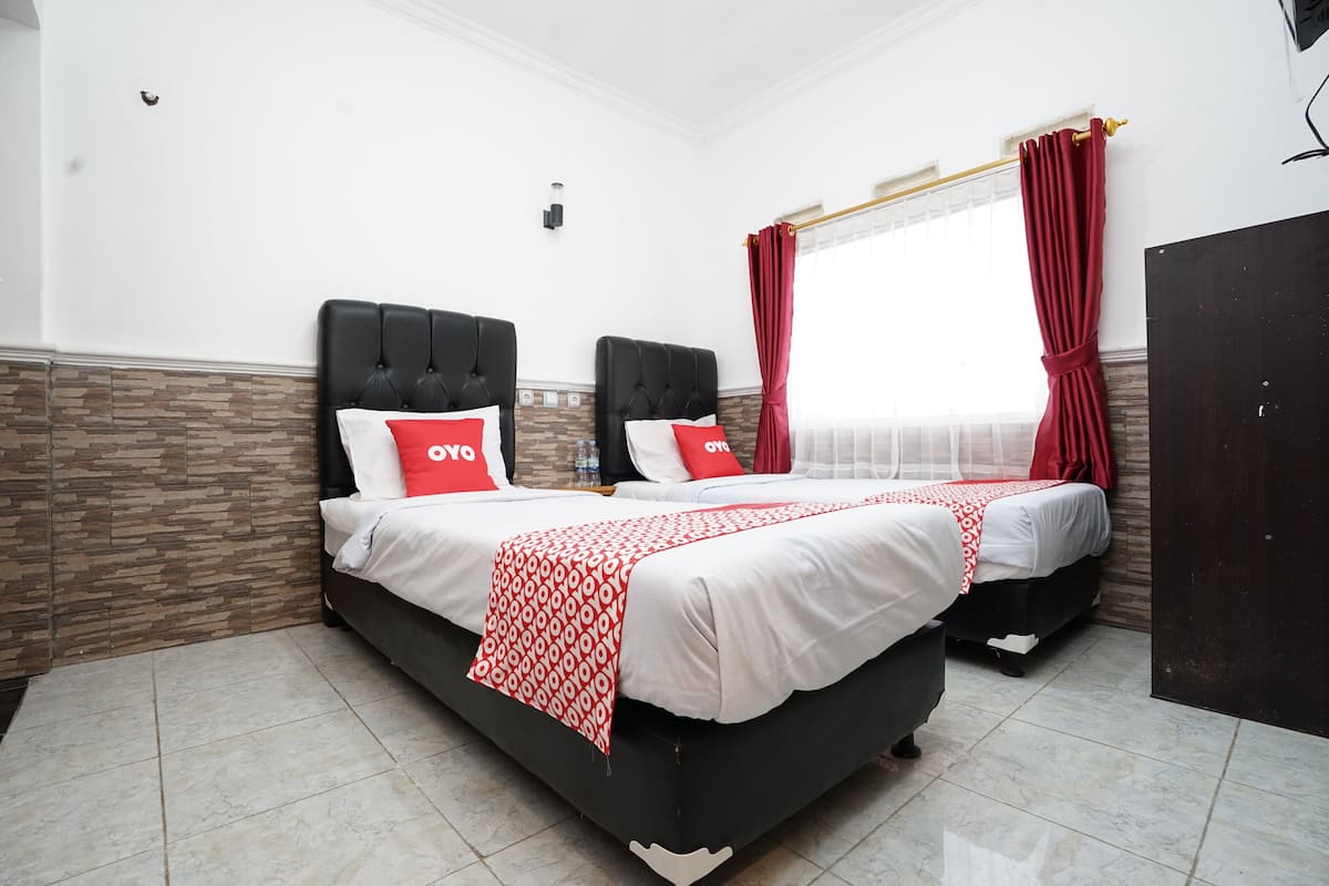 Ein Hotelzimmer mit zwei Betten, jeweils mit einem schwarzen Kopfteil, weißer Bettwäsche und roten Kissen. Der Raum hat einen gefliesten Boden und ein Fenster mit roten Vorhängen.