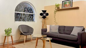 Una sala de estar con un sofá gris oscuro, una mesa de centro redonda de cristal, un sillón gris, una araña, un amplio ventanal de arco con ornamentos de hierro forjado y un cuadro en la pared.