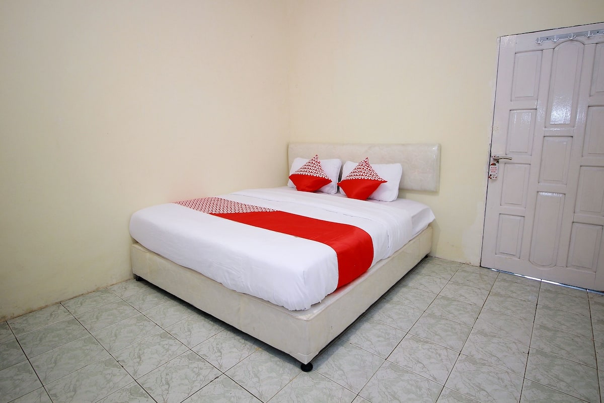 Un lit bien fait, avec une literie blanche et rouge, un mur uni et une porte fermée.