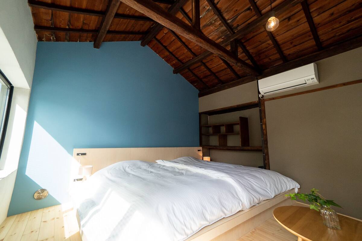 A bedroom with a wooden ceiling, a blue wall, a bed, a bedside table, and a window.