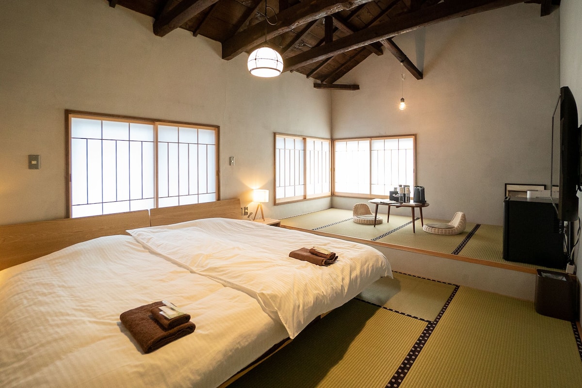 A traditional Japanese-style room with tatami flooring, a large bed with white bedding, a small table with a chair, and a television.