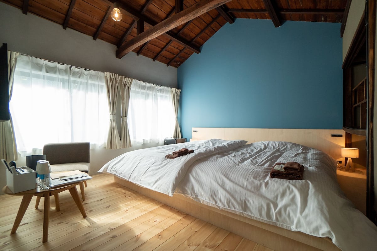 A bedroom with a large bed, wooden floor, a desk with a lamp, and a chair.
