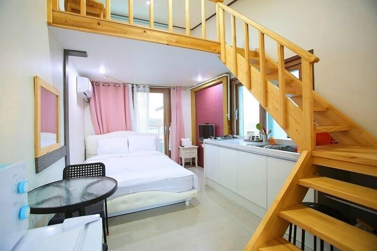 Ein Schlafzimmer mit Bett, einem kleinen Tisch, einem Schreibtisch, einer Küchenzeile und einer Holztreppe.