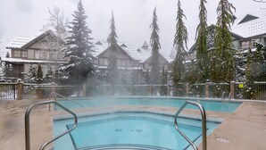 Zona de piscina con barandillas metálicas, rodeada de árboles y edificios cubiertos de nieve.