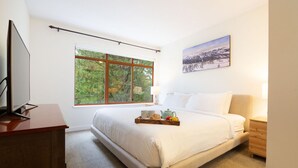 Un dormitorio con una cama grande, un televisor sobre un escritorio de madera, una ventana con vista a árboles y un cuadro en la pared.