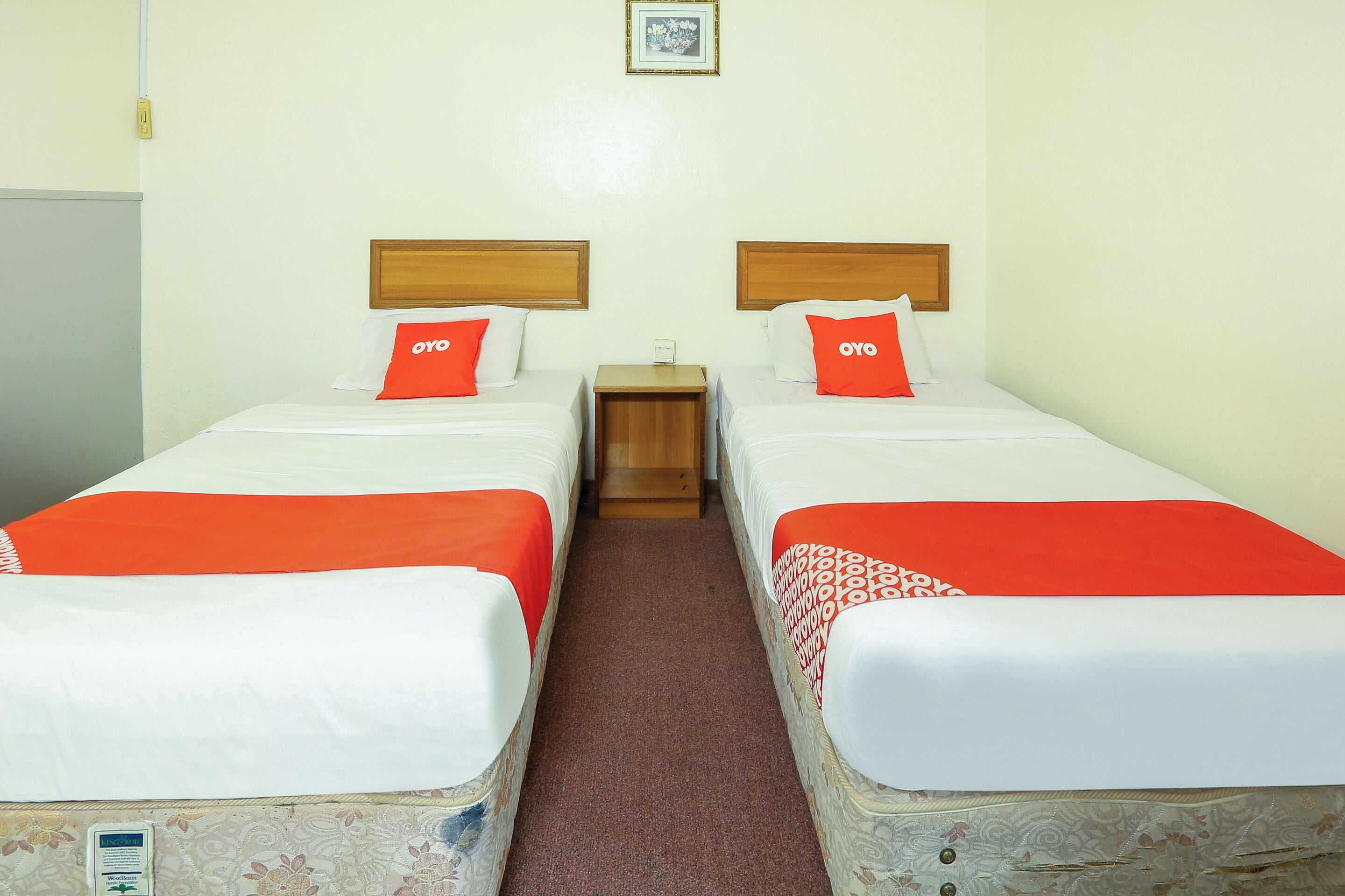 A hotel room with two beds, each with a wooden headboard, white bedding, and red accents. There is a small wooden nightstand between the beds, and a framed picture on the wall above.