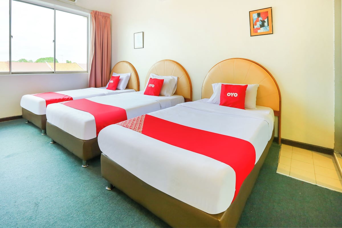 A hotel room with three beds, each with a red and white bedspread and a pillow with "POYO" on it. The room has a large window with curtains, a framed picture on the wall, and a green carpet.