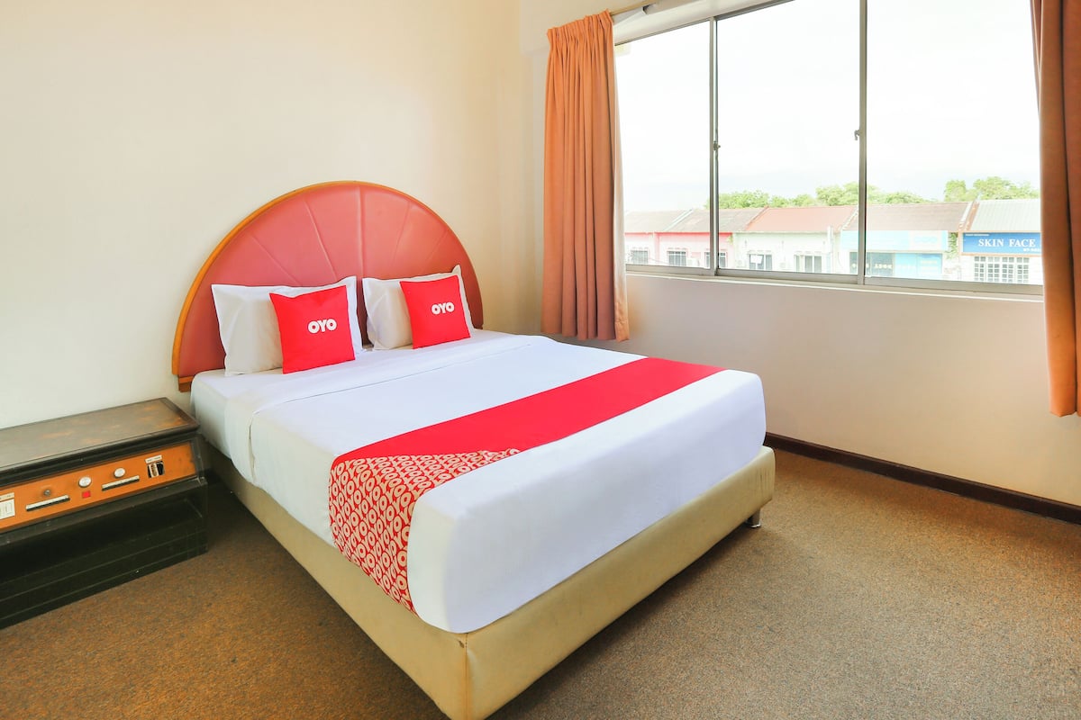 A hotel room with a bed, a nightstand, a window with curtains, and a view of buildings outside.