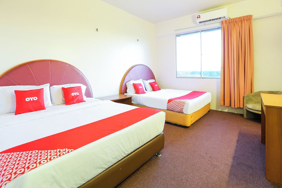 A hotel room with two beds, each with a red headboard and white bedding. There is a window with curtains, a desk, and an air conditioning unit on the wall.