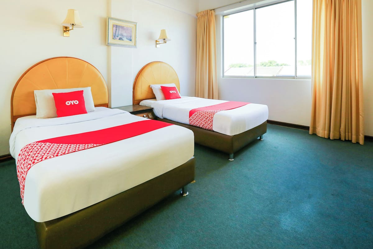 A hotel room with two beds, each with a headboard, a window with curtains, and wall-mounted lamps.