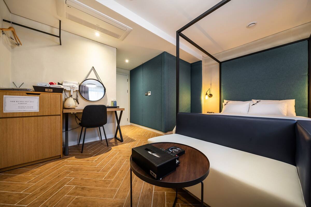 A modern hotel room with a wooden desk, a black chair, a round mirror, a bed with a dark headboard, and a small table with a remote control.