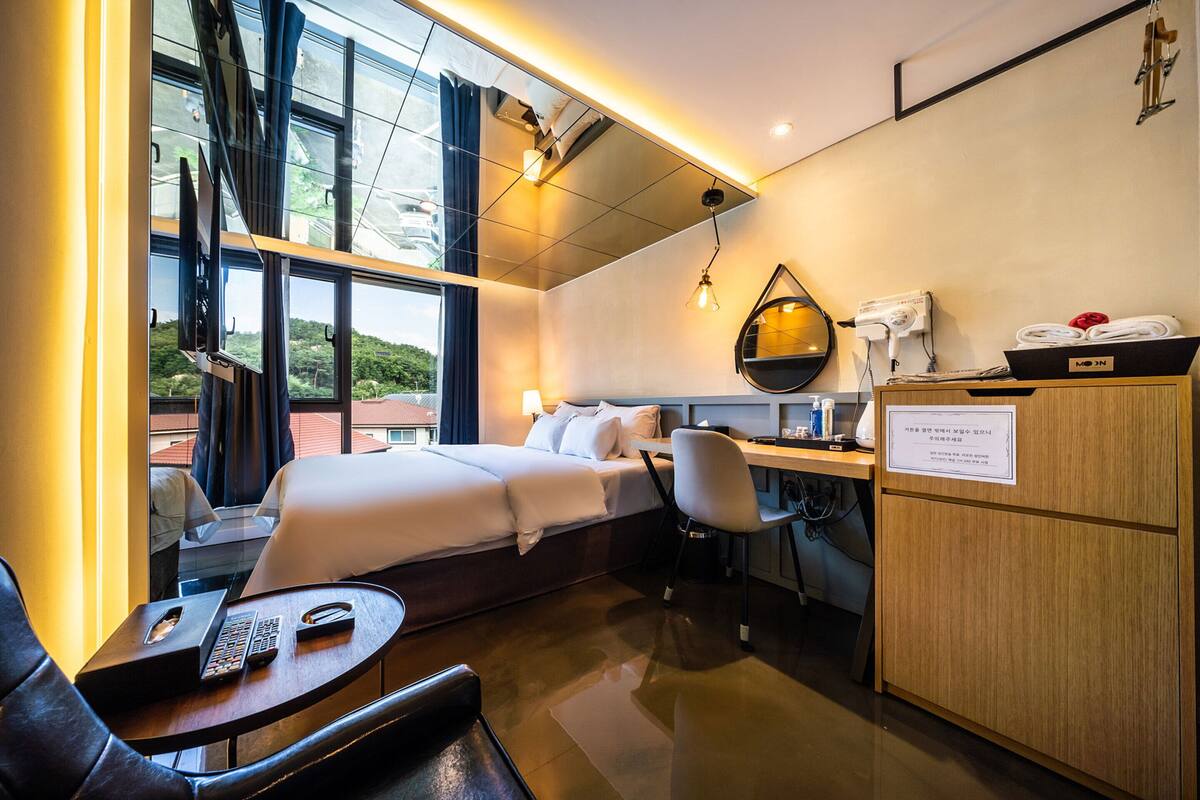A modern hotel room with a large bed, a desk, and a chair. There is a window with a view of buildings and trees.