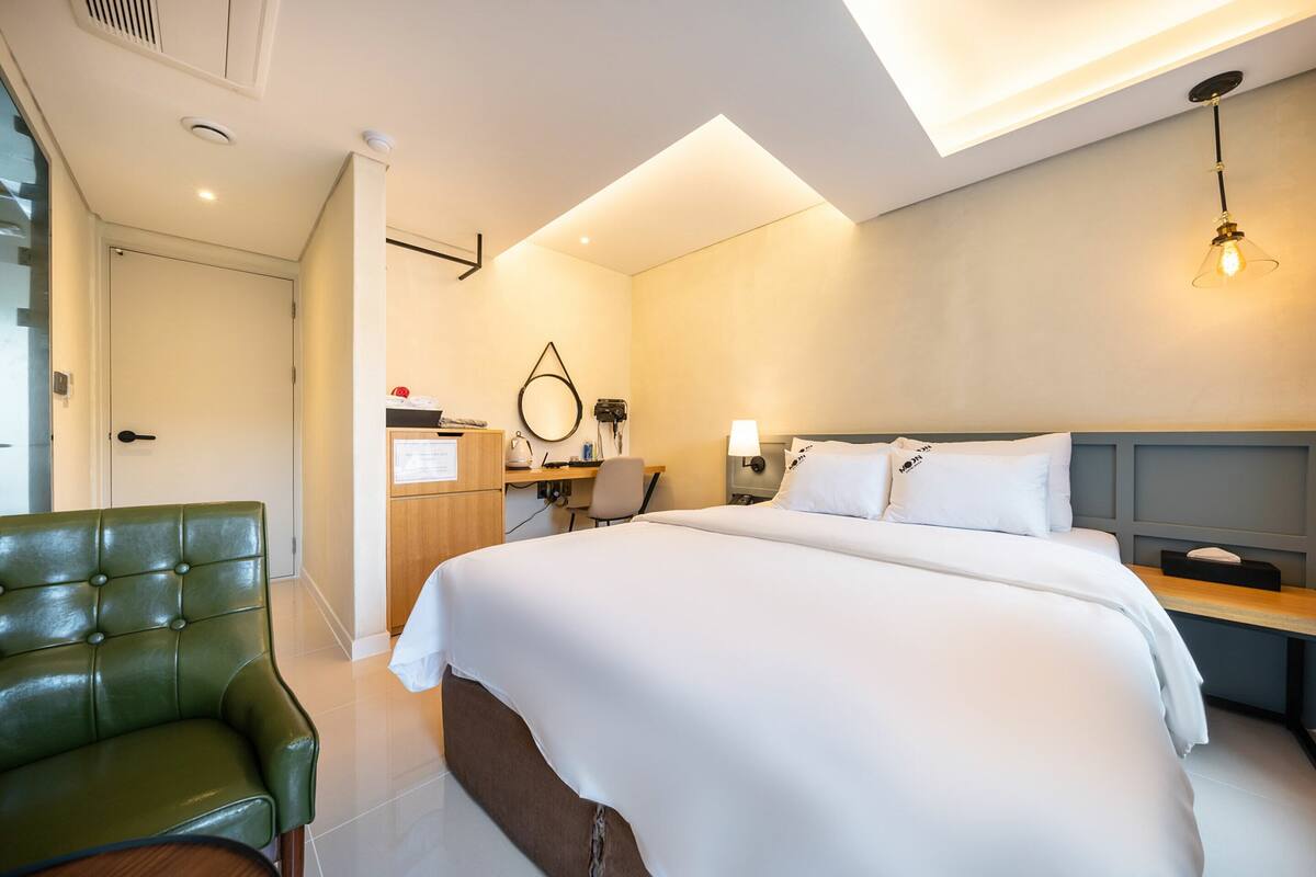 A modern hotel room with a large bed, a green leather chair, a desk with a computer, and a desk lamp.