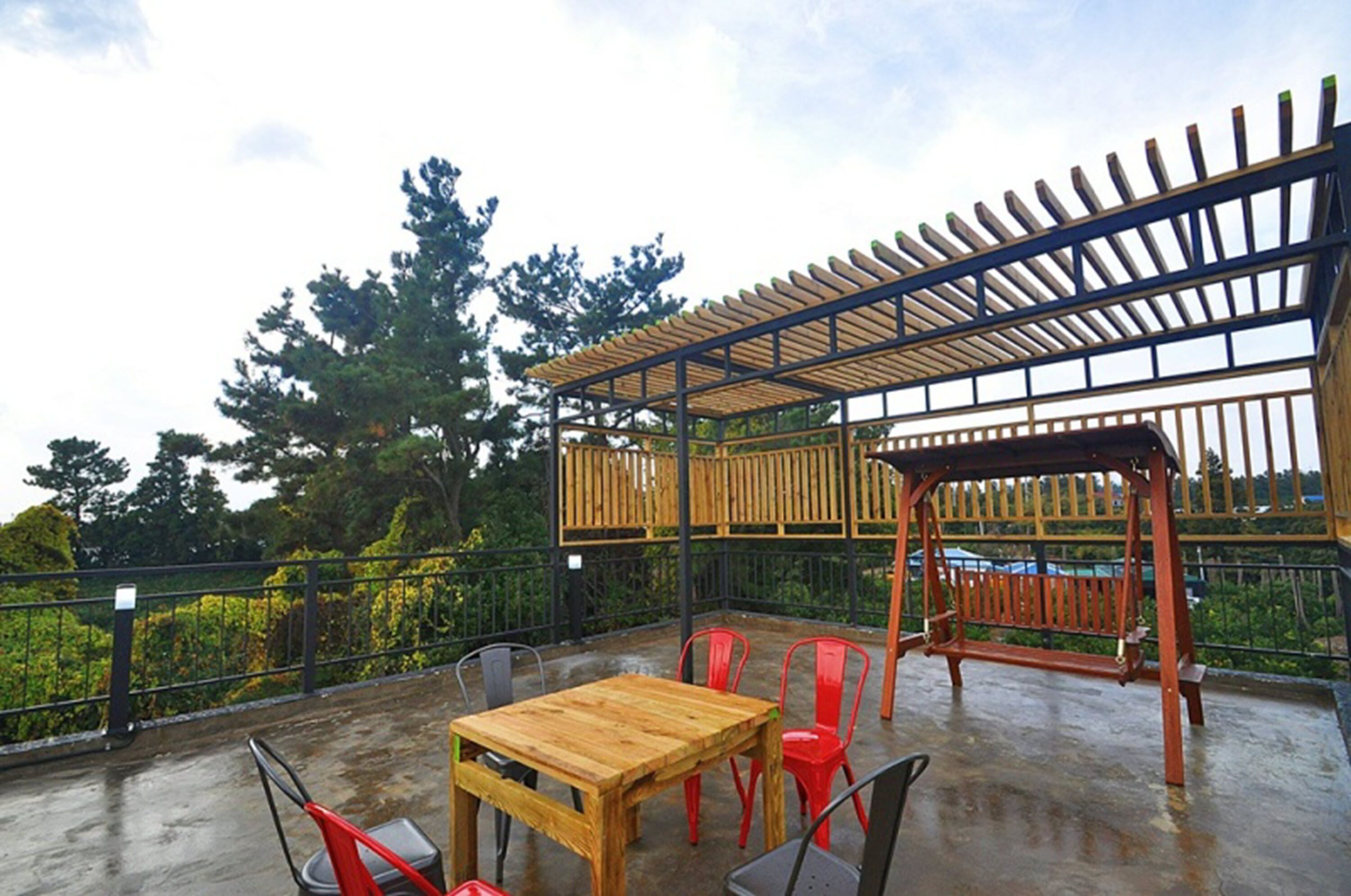 An outdoor patio with a wooden table and chairs, a pergola, and a view of trees and greenery.