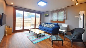 A modern living room with a blue sofa, a glass coffee table, a wicker chair, and a sliding glass door leading to a balcony. - Nonil So Pension (Seogwipo)