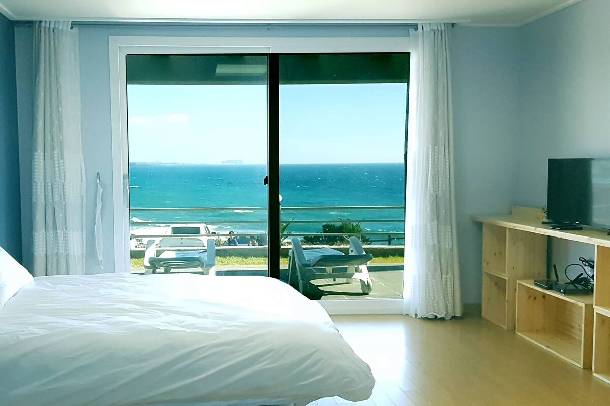Une chambre d’hôtel avec un grand lit, un téléviseur posé sur une étagère en bois et un balcon offrant une vue sur l’océan.