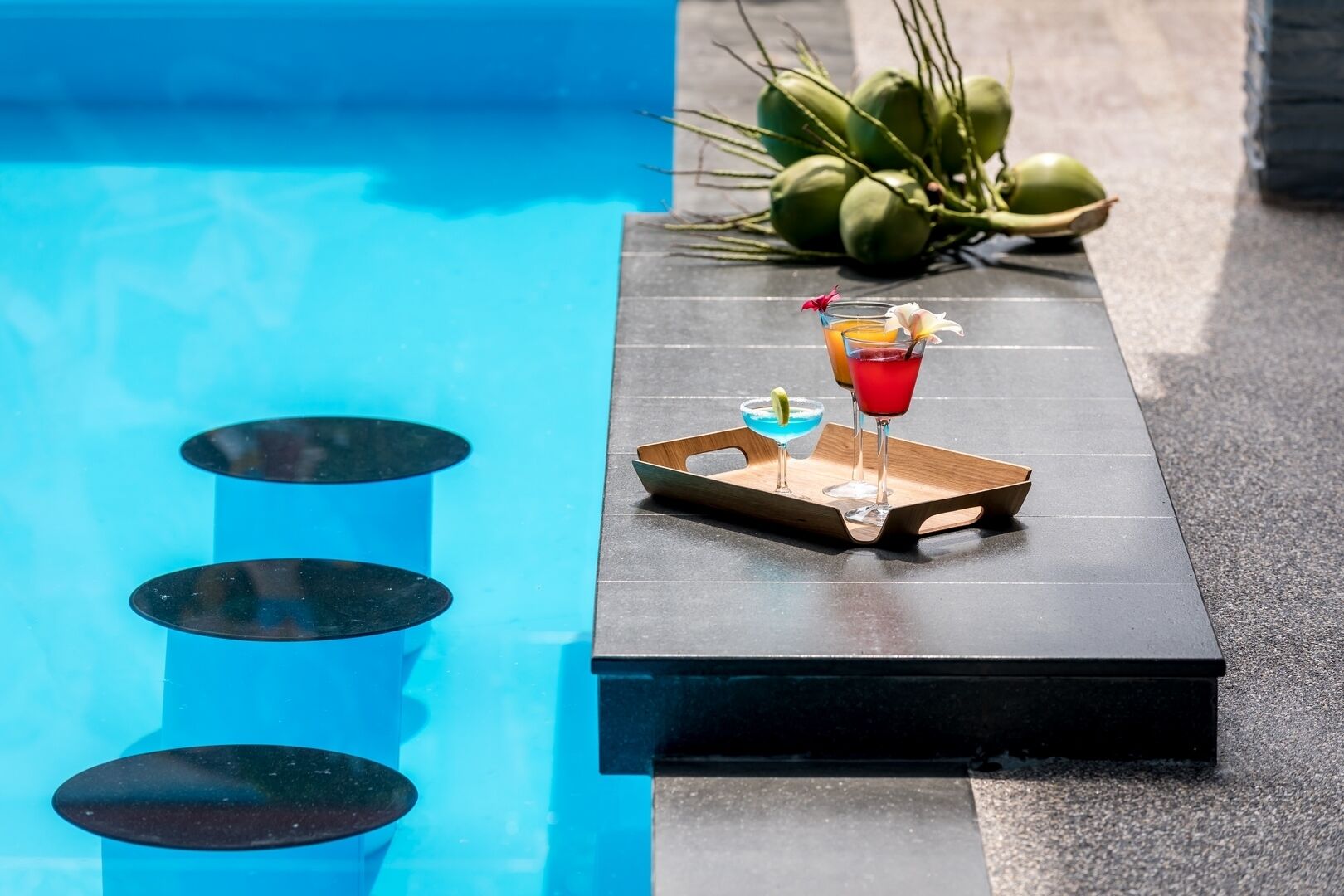 A poolside bar with two drinks on a tray, a pool, and coconuts in the background.