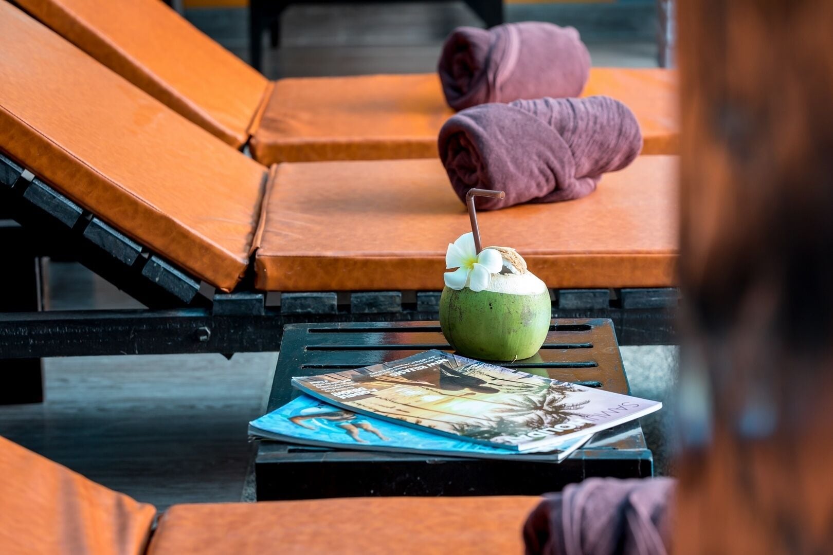A lounge area with orange cushioned chairs, a table with a magazine, and a coconut drink with a straw and flower.