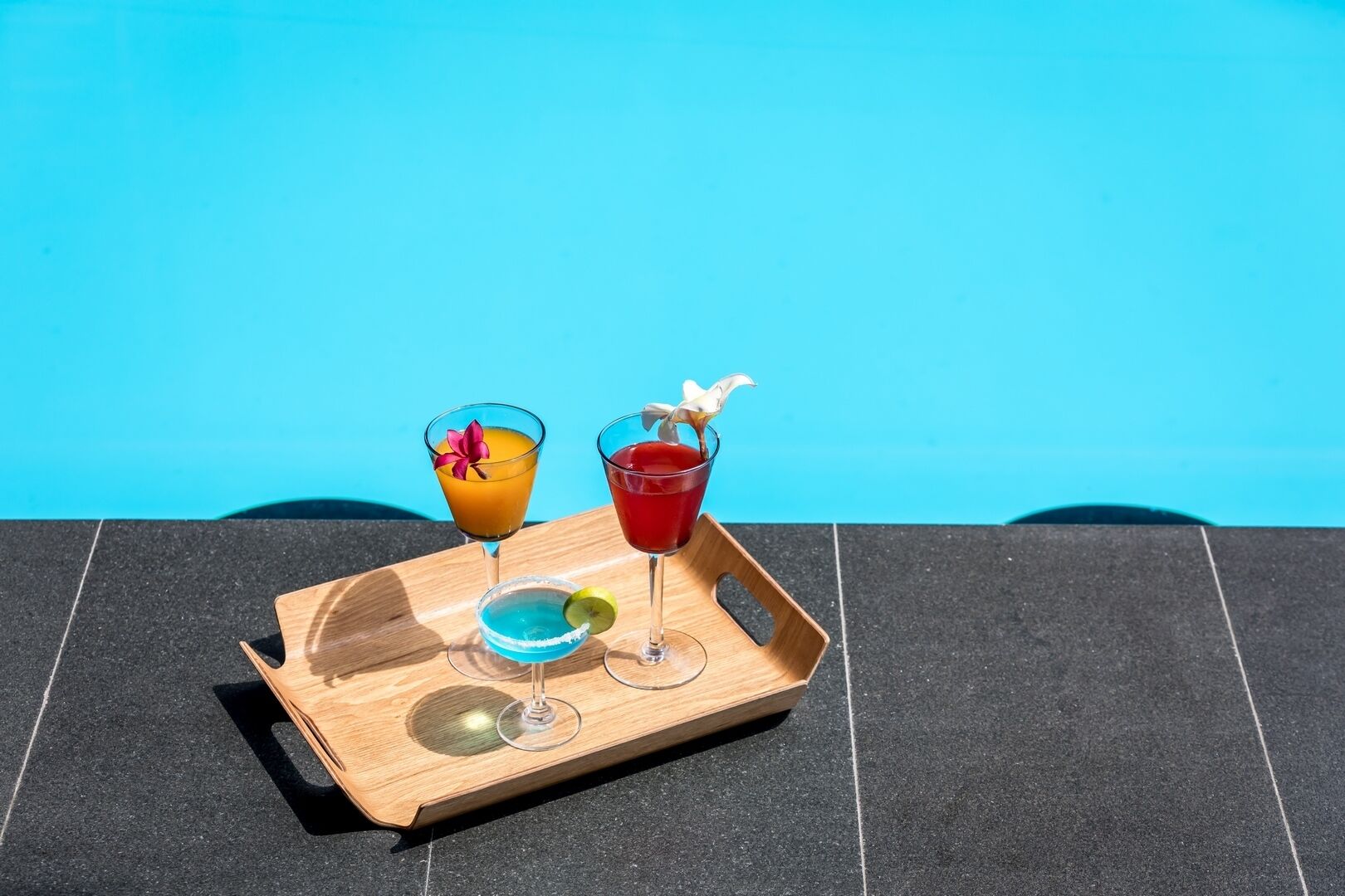 A tray with three drinks and a small bowl on a tiled surface by a pool.