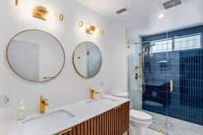 A modern bathroom with two large round mirrors, a white marble sink, a gold faucet, and a glass-enclosed shower area. - Belding Bliss (Palm Springs)