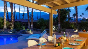 An outdoor dining area with a pool, wooden tables, and chairs, surrounded by palm trees and a mountainous backdrop. - Modern Loma Vista (Palm Springs)