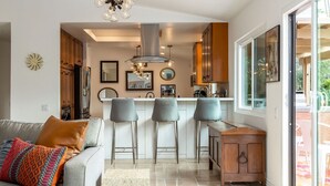 A modern kitchen with a central island, bar stools, and a dining area with a glass door leading to an outdoor space. - Modern Loma Vista (Palm Springs)