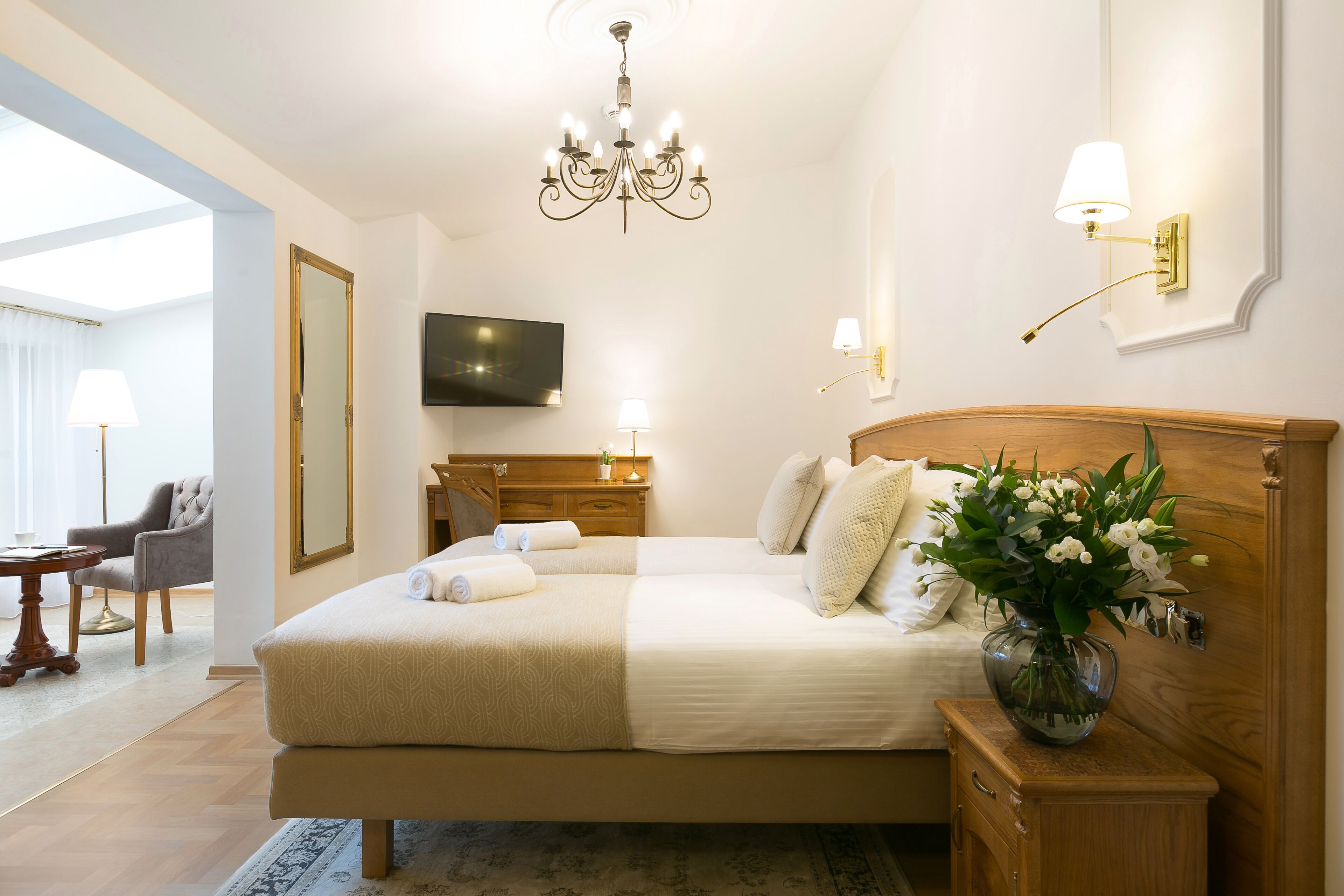 A well-lit bedroom with a large bed, bedside lamps, a TV, and a vase of flowers.