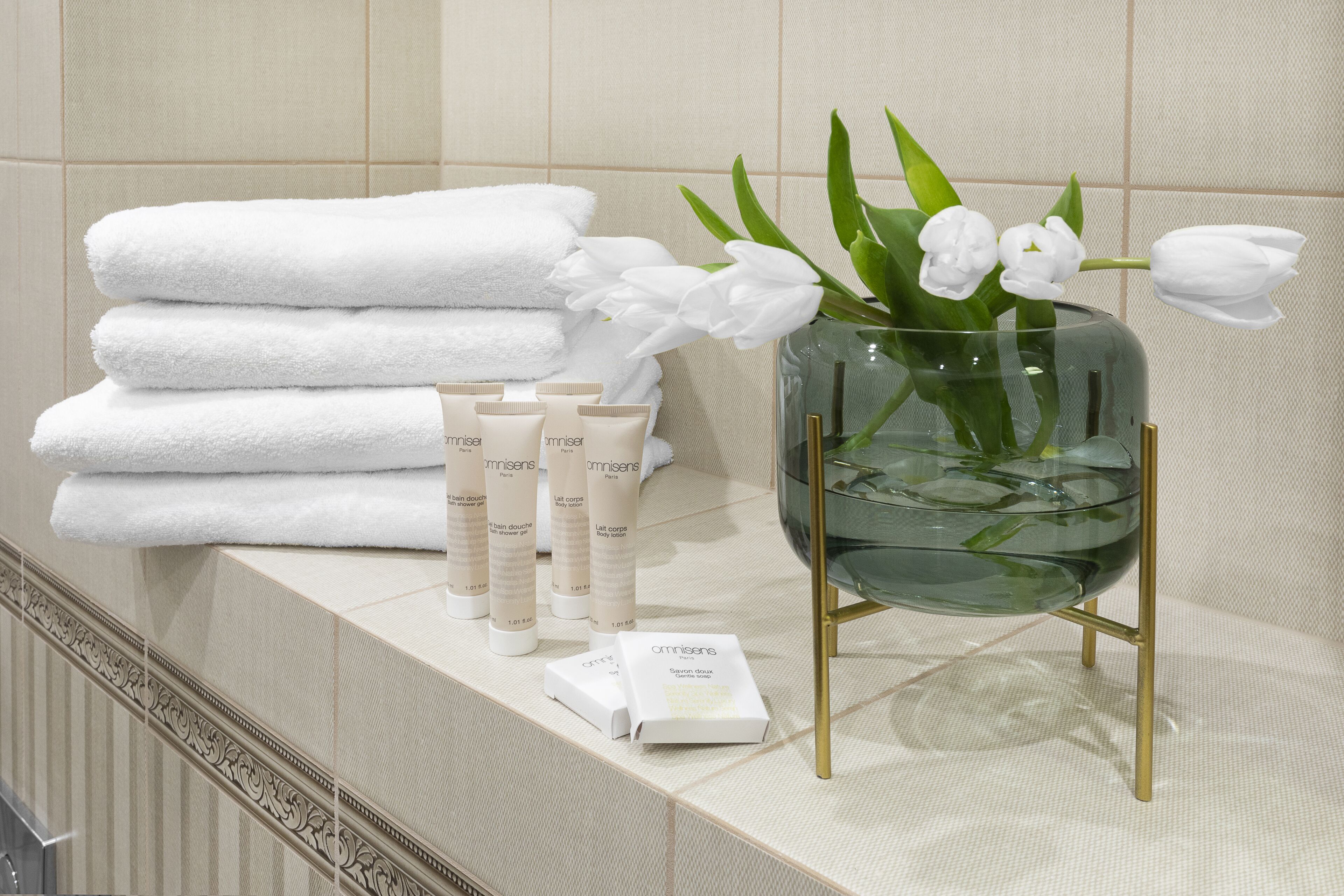 Un baño con toallas blancas ordenadas, un jarrón con flores blancas y artículos de tocador en un estante.