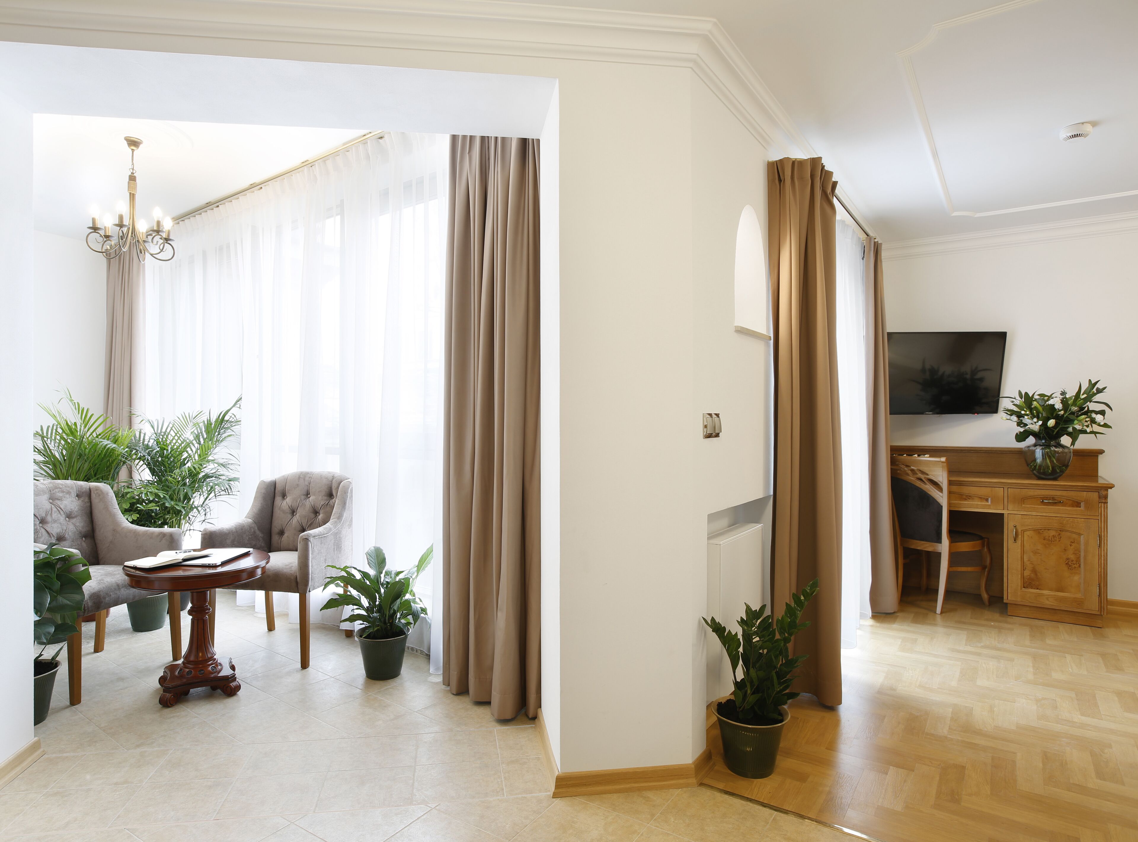 Un salon moderne avec un canapé, des fauteuils, une table basse et des plantes en pot. On y trouve un bureau en bois avec une chaise, et un téléviseur fixé au mur.